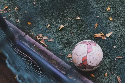 Fußball am Torpfosten auf dunkelgrünem Kunstrasen, ringsum verwehtes Herbstlaub